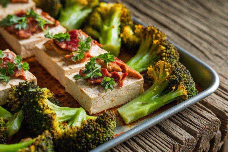 Golden crispy tofu cubes and charred broccoli florets on a parchment-lined sheet pan drizzled with sesame sauce
